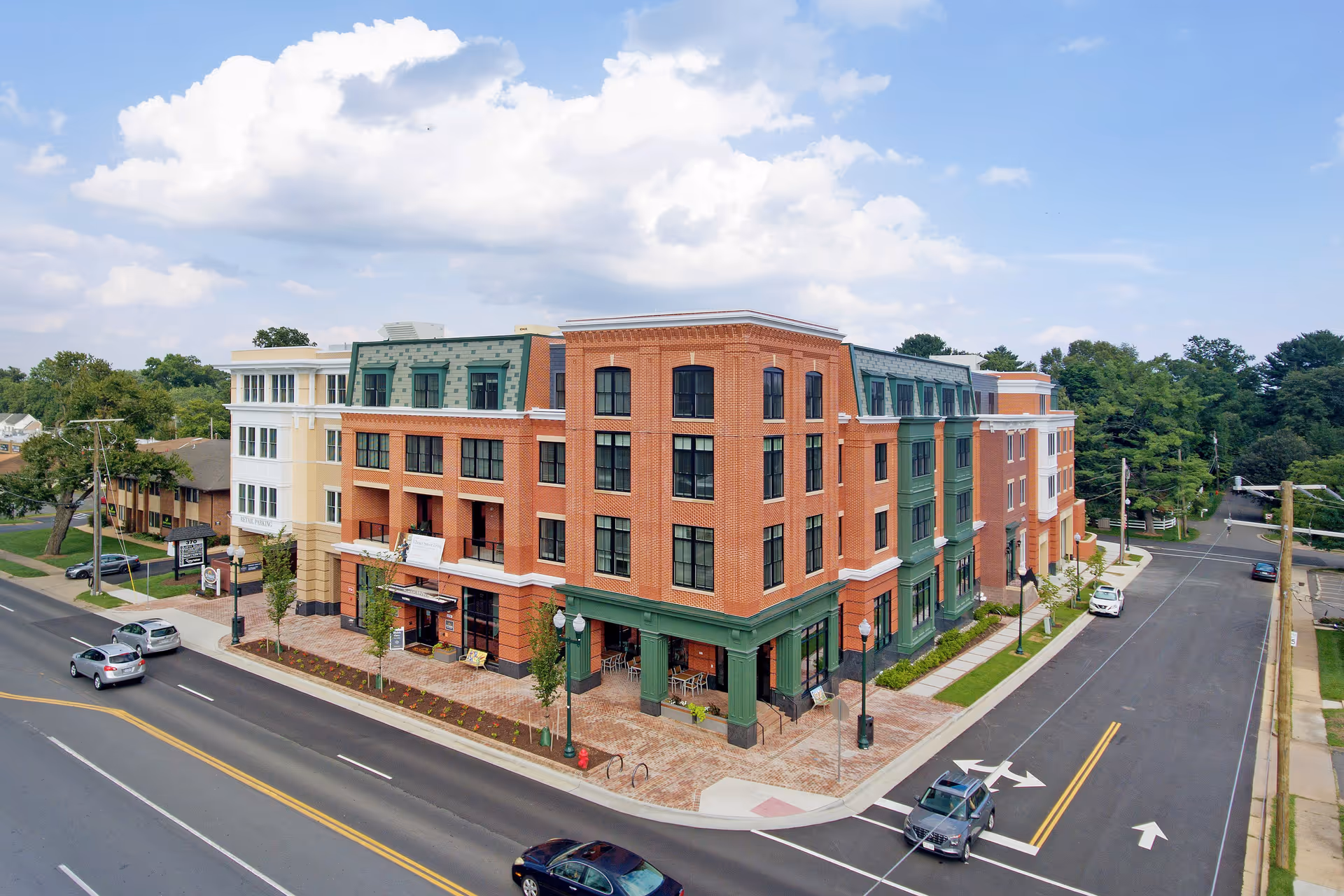 A large, multi-story brick building with green and beige accents situated at a street corner under a partly cloudy sky. Several cars are visible on the roads surrounding the building, which has large windows and outdoor seating areas on the ground floor.