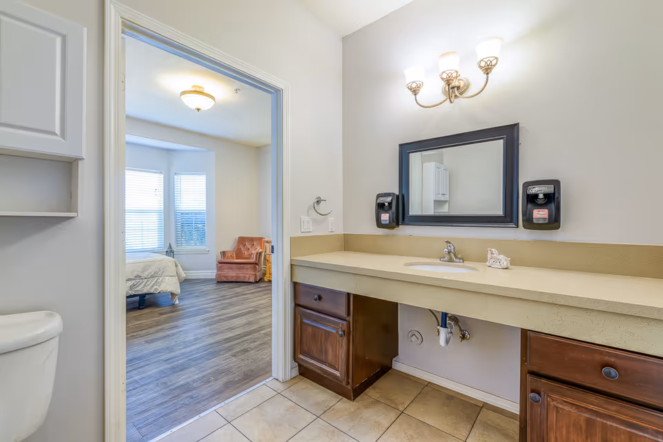 View of a bathroom with a long countertop and sink, a mirror with two soap dispensers on either side, and a light fixture above. The bathroom opens into a bedroom with a bed, a pink armchair, and windows with blinds.