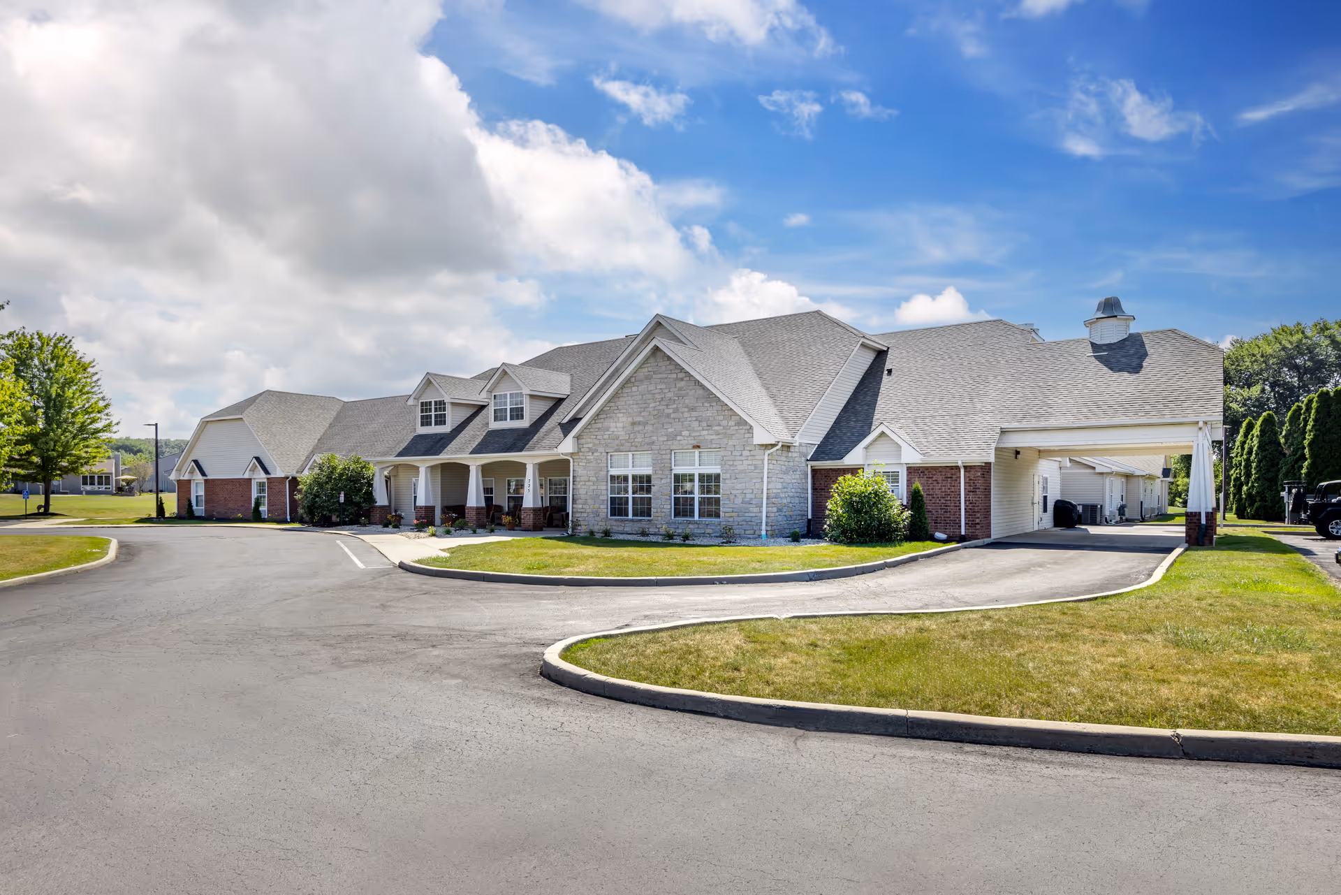 Front exterior of a single-story senior living facility with a covered entrance, circular driveway, and landscaped lawn under a partly cloudy sky.