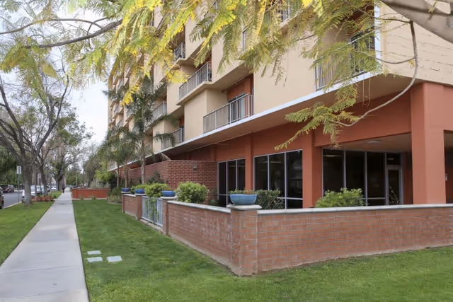 Exterior view of a multi-story residential building with balconies, a brick and stucco facade, and a small enclosed garden area with potted plants. A sidewalk runs alongside the building with trees lining the street.