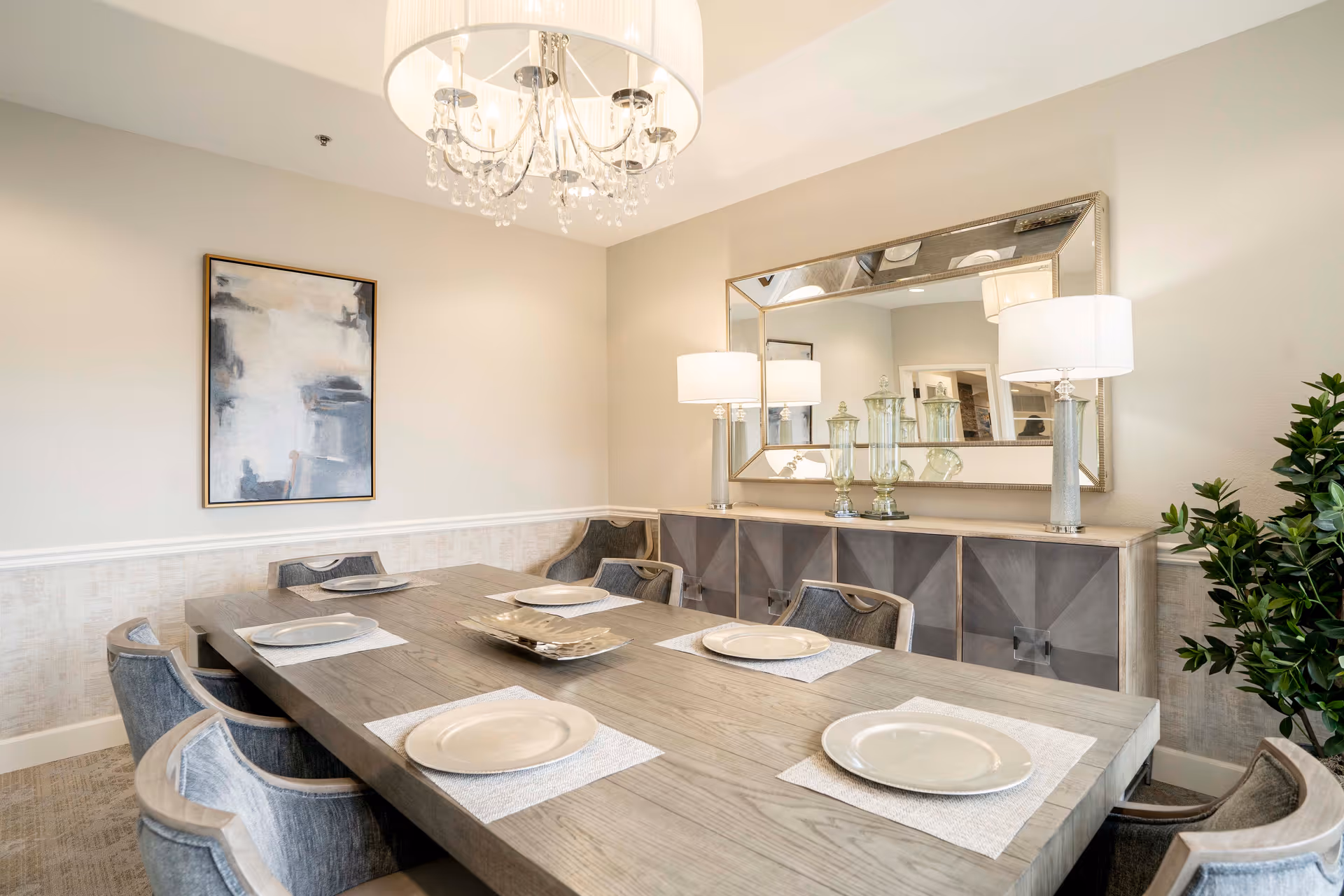 A modern dining room with a large wooden table set with six plates on white placemats. Six upholstered chairs surround the table. A large mirror hangs on the wall above a sideboard with decorative glass jars and two table lamps. A chandelier with crystal accents hangs from the ceiling, and a framed abstract painting is on the adjacent wall. A green plant is visible in the corner.