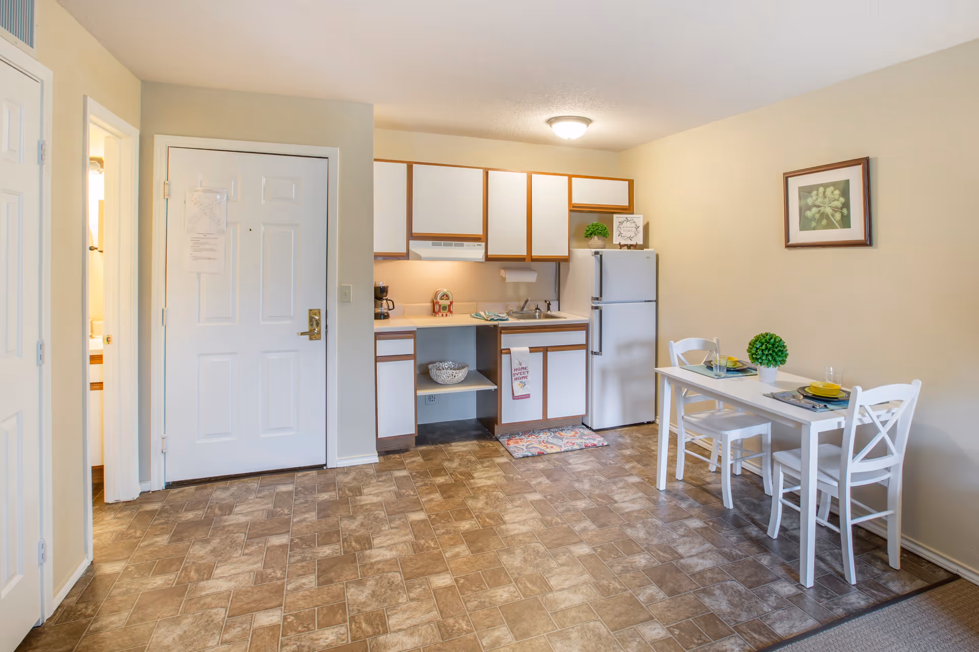 A small kitchen area in a senior living facility with white cabinets and a white refrigerator. There is a small dining table with two white chairs, set with plates, glasses, and a small green plant centerpiece. The floor has a brown patterned tile, and there is a framed picture on the beige wall. A door and a bathroom entrance are visible on the left side.