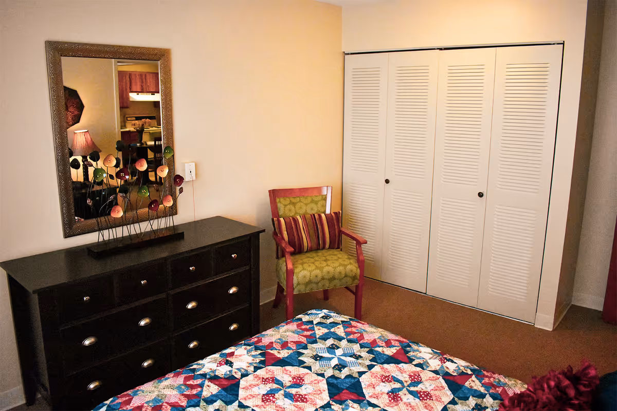 A bedroom with a colorful quilted bedspread, a green armchair with a striped cushion, a dark wooden dresser with a decorative flower sculpture on top, and a large mirror hanging above the dresser. There is a white closet with louvered doors on the right side of the room.