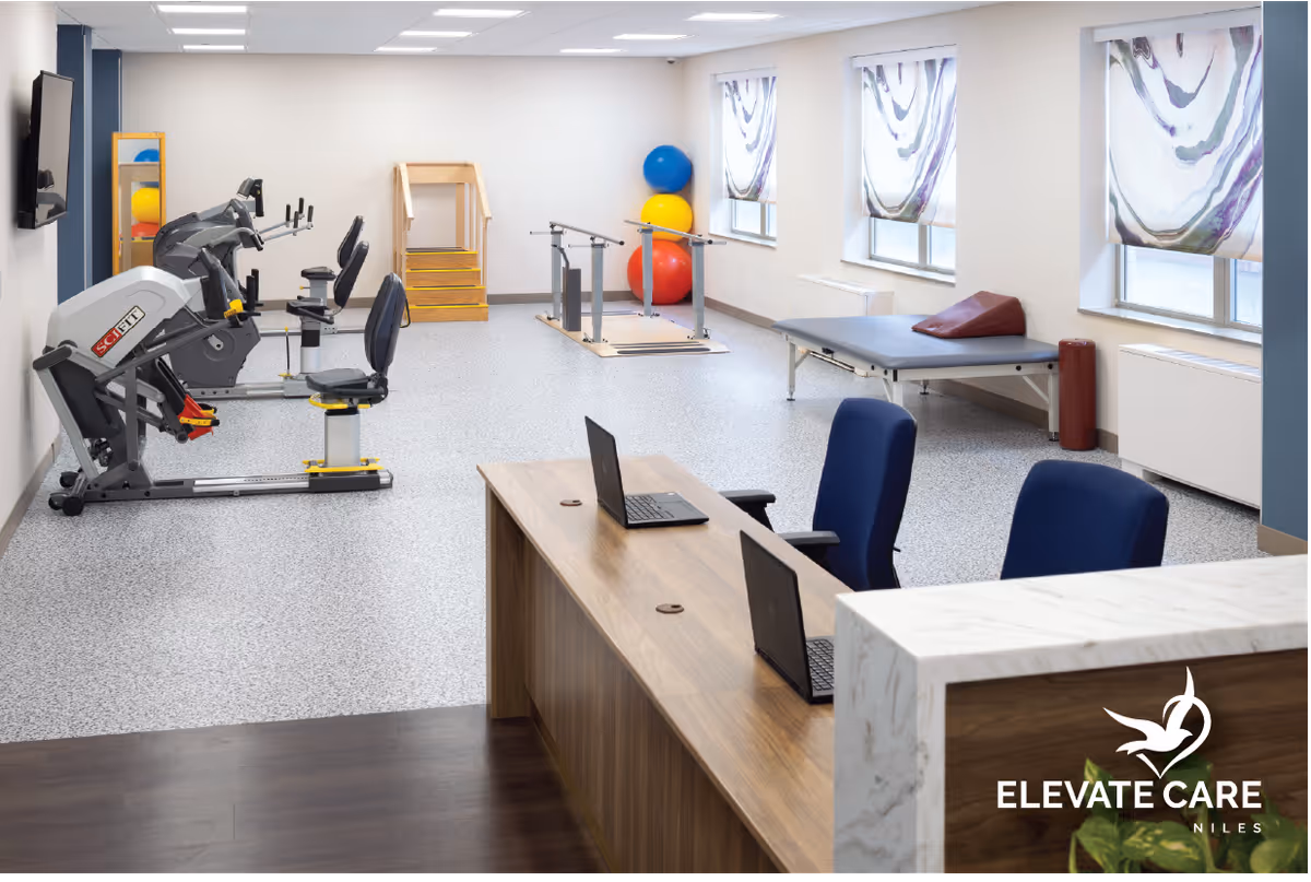 A physical therapy or rehabilitation room with exercise equipment including stationary bikes, parallel bars, therapy balls, a therapy table with a wedge cushion, and a wooden step platform. In the foreground, there is a reception desk with two laptops and two blue chairs. The room has large windows with patterned curtains and bright overhead lighting.