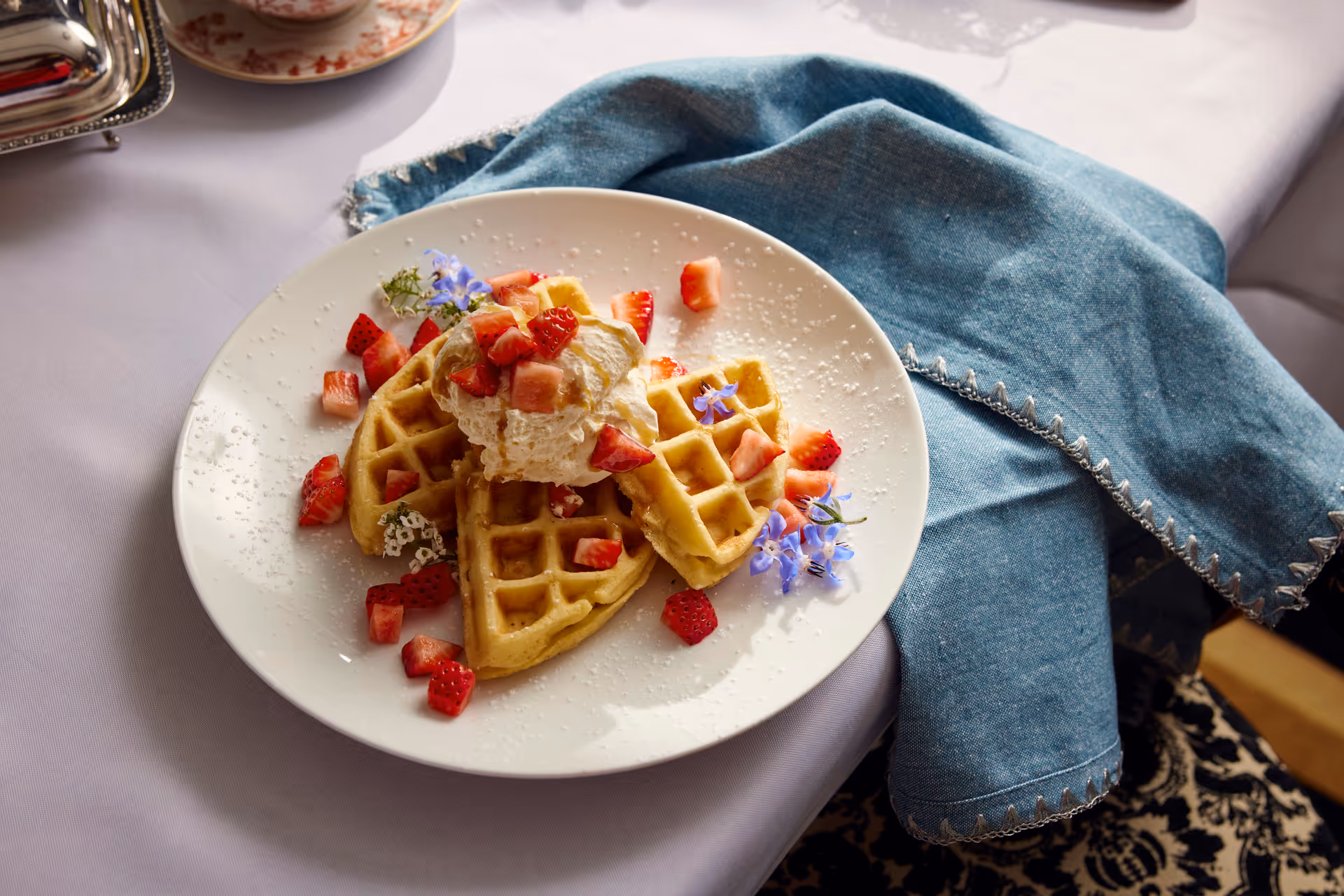 A white plate with three heart-shaped waffles topped with whipped cream, diced strawberries, and small purple edible flowers, placed on a white tablecloth with a blue cloth napkin beside it.
