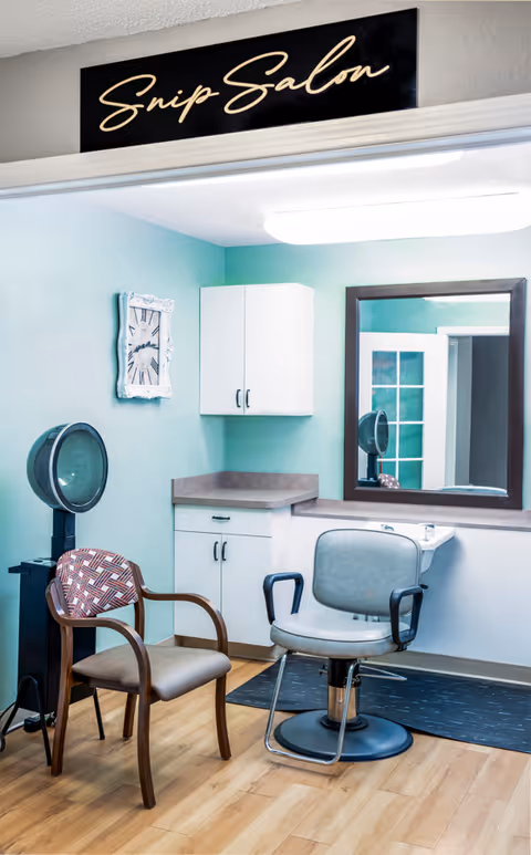 Interior view of a salon room with a sign above the door reading 'Snip Salon'. The room features a hair drying station with a vintage hooded dryer, a wooden chair with a patterned cushion, a salon chair in front of a large mirror, white cabinets, and a wall clock with a decorative frame. The walls are painted light teal and the floor is light wood.