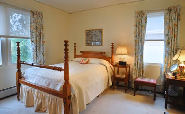 A traditional furnished bedroom with a wooden four-poster bed, bedside tables with lamps, floral curtains and windows.