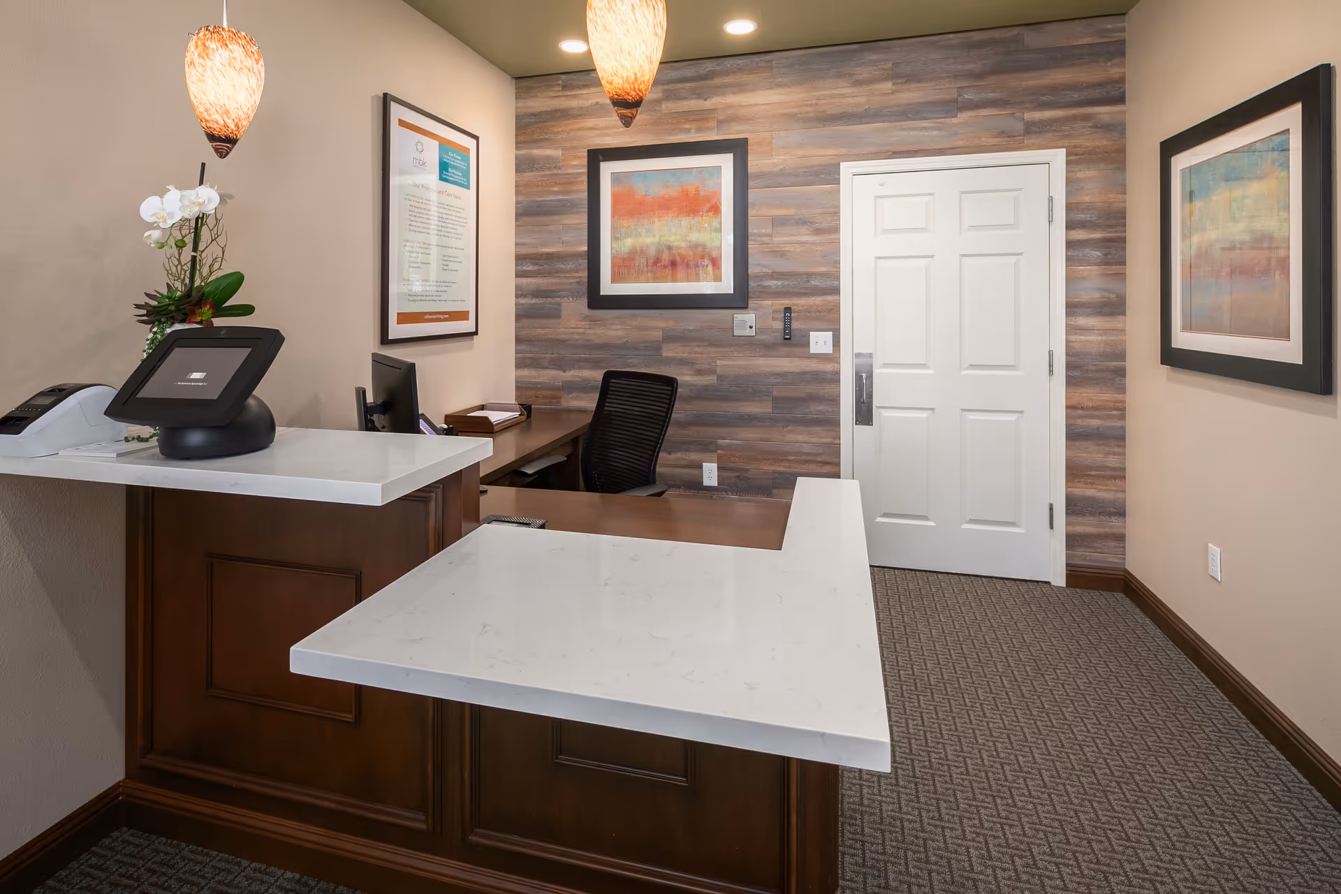 Reception desk area with a white countertop and dark wood base, a computer monitor, a phone, and a small plant with white flowers. Behind the desk is a black office chair and a wooden wall with two framed abstract paintings. A white door is visible on the right side of the wall. Two pendant lights hang from the ceiling.