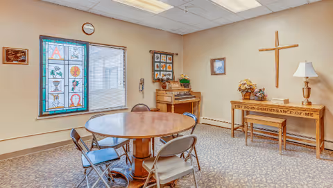 A small room with a round wooden table surrounded by four folding chairs. The room has a stained glass window with religious symbols, a wall clock, a wooden organ, and a wooden table with a cross mounted above it. The table has a lamp and a floral arrangement, and the wall behind it has a framed picture. The carpet has a patterned design.