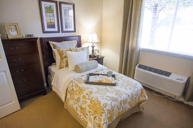 A cozy bedroom with a single bed dressed in floral patterned bedding and multiple pillows. Next to the bed is a nightstand with a lamp and a small decorative item. A tall wooden dresser stands against the wall, and two framed pictures hang above the bed. A large window with curtains lets in natural light, and an air conditioning unit is installed below the window.