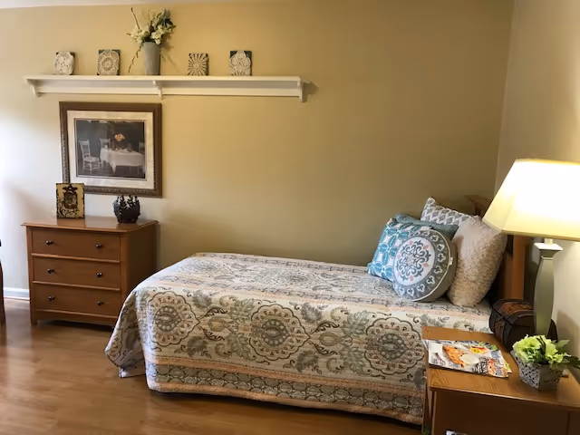A cozy bedroom with a single bed covered in a patterned quilt and multiple decorative pillows. Next to the bed is a wooden nightstand with a lamp, a small plant, and some magazines. On the opposite side of the room is a wooden dresser with three drawers, decorated with framed artwork and a vase with flowers. Above the dresser is a white wall shelf holding decorative items. The walls are painted a warm beige color and the floor is wooden.
