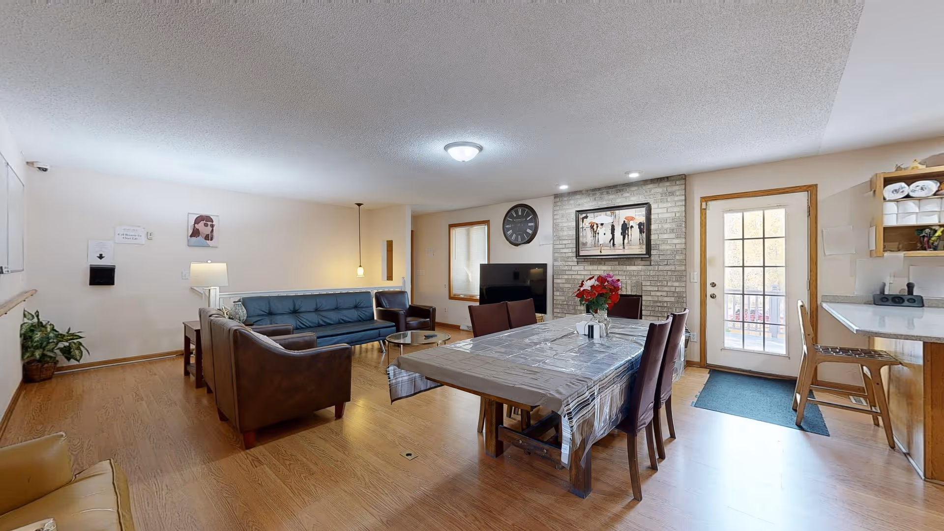 A spacious, well-lit common area with wooden flooring featuring a dining table covered with a plastic tablecloth and six chairs. To the left, there is a seating area with a blue couch, two brown armchairs, and a small round coffee table. The back wall has a large clock, a framed picture, and a TV on a stand. A door with glass panes leads outside, and there is a kitchen counter with two chairs on the right side. The room has a cozy and welcoming atmosphere.