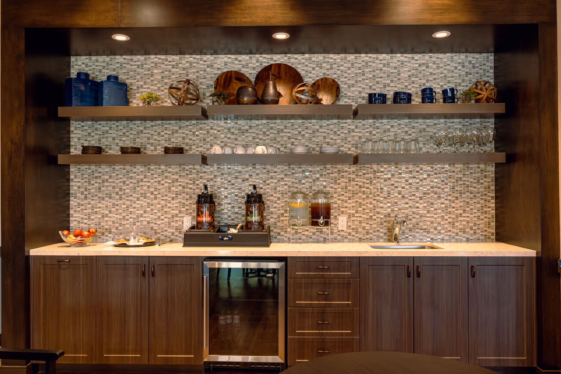 A small kitchenette/bar area with a mosaic tile backsplash, floating shelves with dishes and decor, beverage dispensers, a sink and dark wooden cabinets.