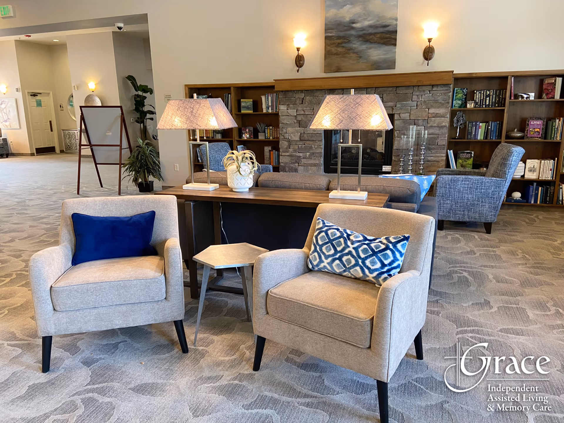 A cozy living room area in an assisted living facility featuring two beige armchairs with decorative pillows, a wooden table with two lamps and a plant, a stone fireplace with a painting above it, and bookshelves filled with books in the background.