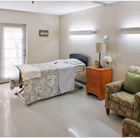 A hospital-style bedroom with bed, nightstand, lamp, and armchair in a neutral room.