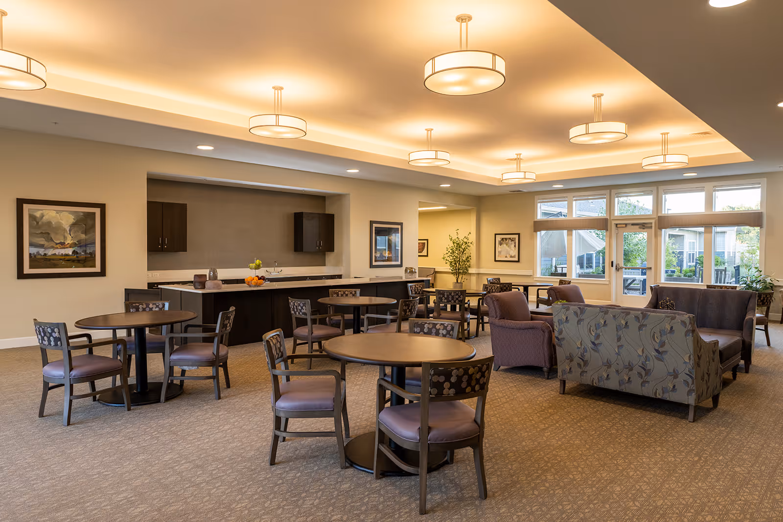 A spacious and well-lit common area in a senior living facility with several round tables and chairs arranged for seating. There are also cushioned armchairs and sofas with floral patterns. The room features large windows allowing natural light to enter, framed artwork on the walls, and modern ceiling lights. A counter with cabinets is visible in the background, along with some decorative items like fruit bowls and plants.