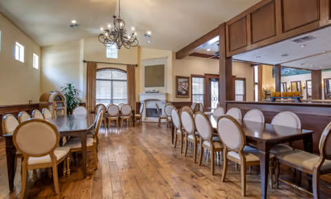 Spacious dining room with wooden floors, multiple tables and chairs arranged neatly, a chandelier hanging from the ceiling, large windows with curtains, a fireplace, and a jukebox in the corner.