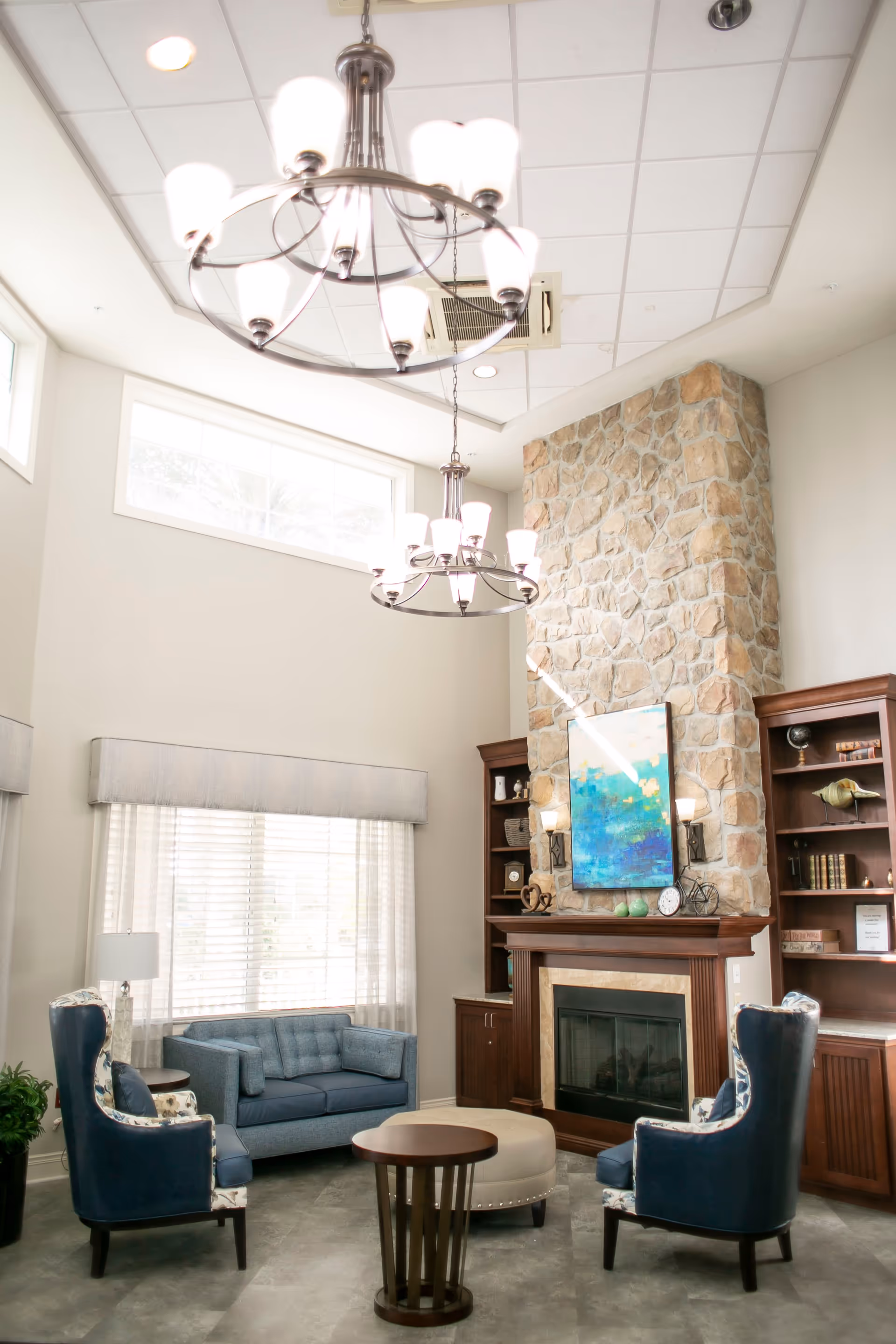 Bright high-ceilinged sitting area with chandeliers, a stone fireplace, blue upholstered chairs and a sofa arranged around a coffee table.