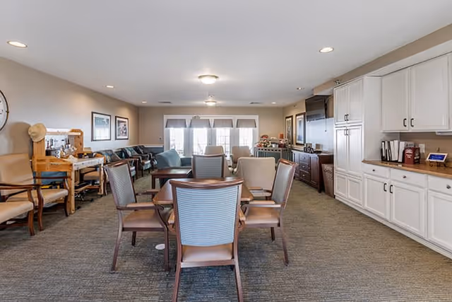 A spacious and well-lit common area with a table and six chairs in the center, surrounded by additional seating including armchairs and sofas. The room features white cabinetry along one wall, framed artwork, and large windows with blinds letting in natural light.