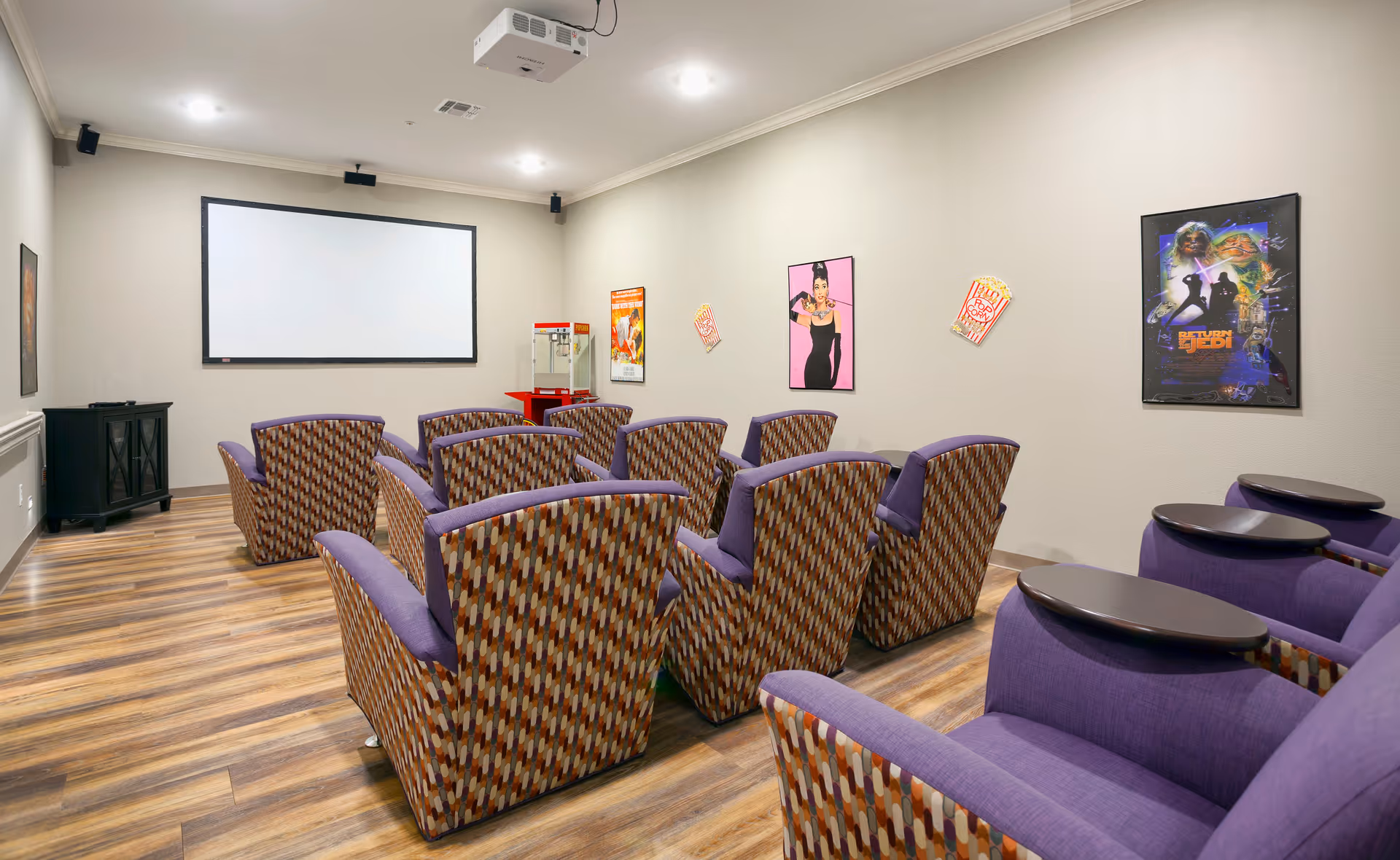 A small movie theater room with two rows of patterned armchairs and a row of purple armchairs with attached dark wood trays. A large white projection screen is mounted on the front wall, with a projector on the ceiling. Movie posters and popcorn decorations are on the walls, and a popcorn machine is in the corner.