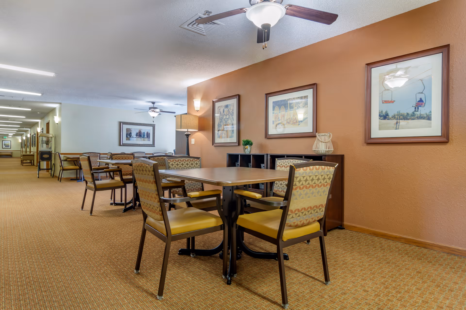 Interior view of a senior living facility common area with several tables and chairs arranged for seating. The walls are painted in warm tones and decorated with framed artwork. Ceiling fans and lamps provide lighting, and a long hallway extends into the background.