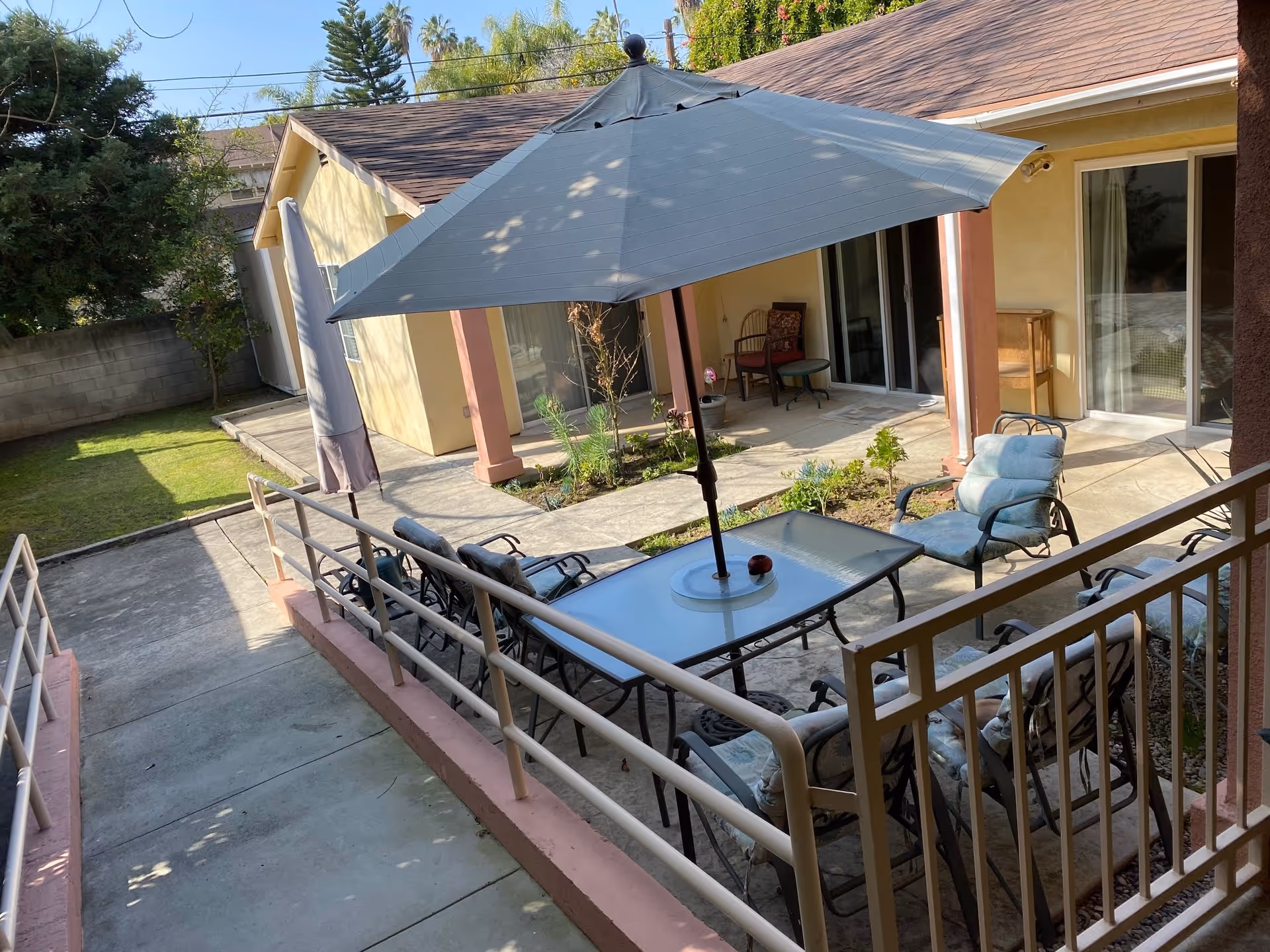 Outdoor patio area at Sunrise Assisted Care Senior Living in Sherman Oaks featuring a glass table with a large umbrella, surrounded by cushioned chairs. The patio is adjacent to a building with sliding glass doors and a small garden area. There is a concrete walkway with handrails leading to the patio.