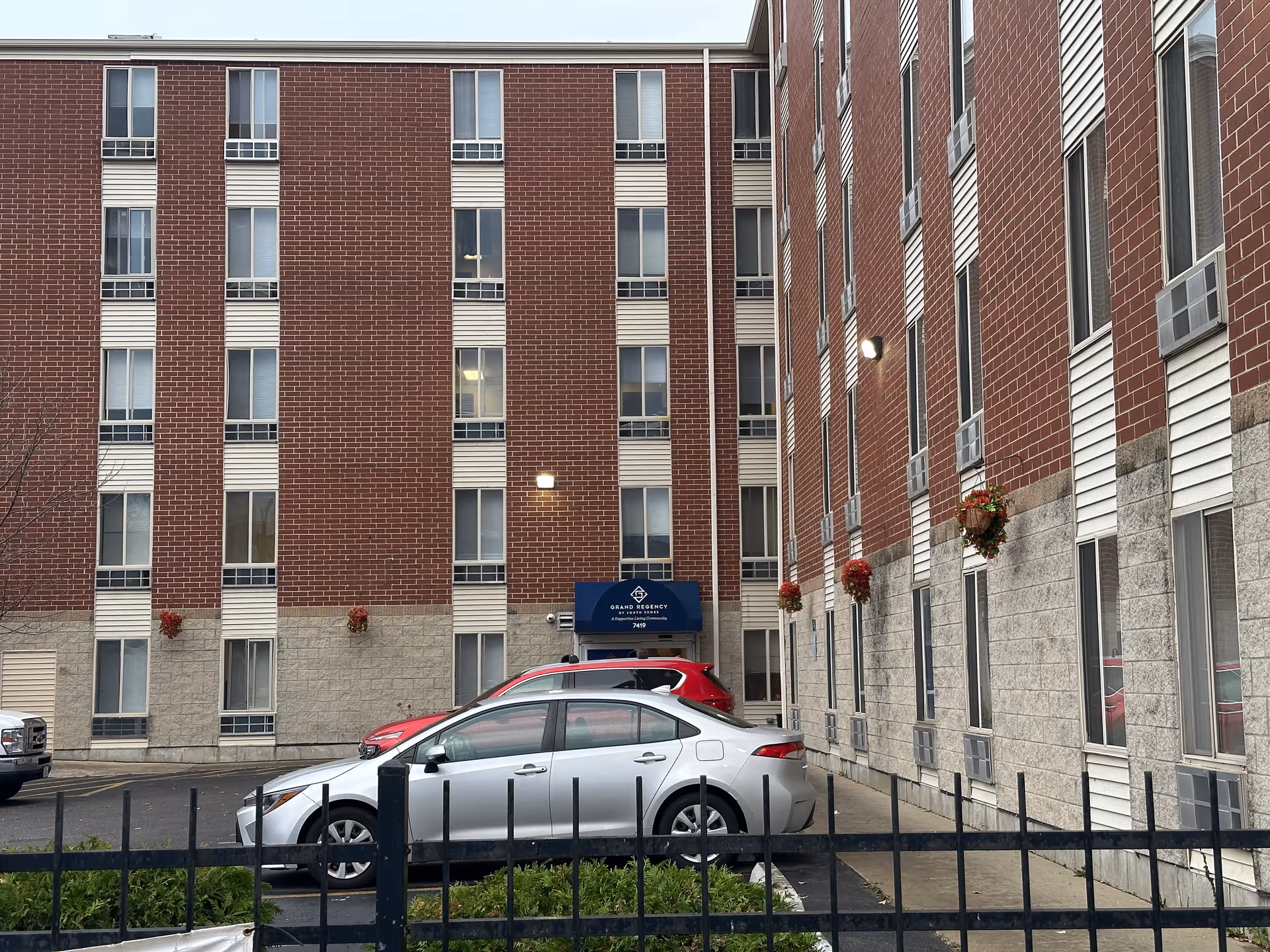 Exterior view of a multi-story brick and stone building with several windows and air conditioning units. There are two parked cars in front of the building, a silver sedan and a red SUV. A blue awning above a door reads 'Grand Regency A Supportive Community 7419'. Flower baskets hang on the wall near the entrance. A black metal fence and some greenery are visible in the foreground.