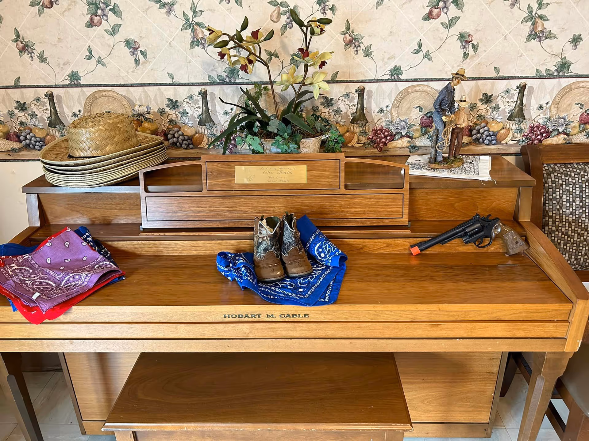 A wooden piano with various decorative items on top including a stack of straw hats, a potted plant with yellow flowers, a figurine of two cowboys, a toy revolver with an orange tip, several colorful bandanas, and a pair of small cowboy boots. The piano has a plaque that reads 'In Loving Memory of Edna Martin You Live on in our Hearts' and the name Hobart M. Cable on the front. The background features wallpaper with a grapevine and fruit pattern.