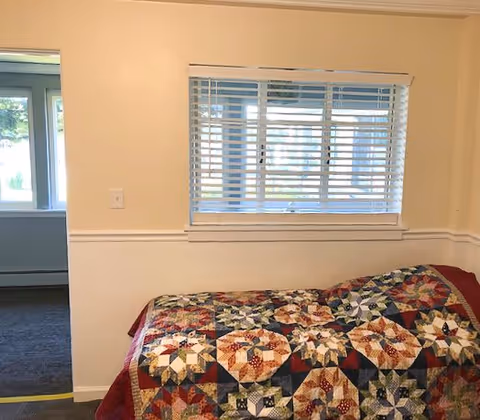 A small bedroom with a bed covered in a colorful patchwork quilt featuring star patterns. The room has beige walls, a window with white blinds, and a doorway leading to another room with a window and carpeted floor.