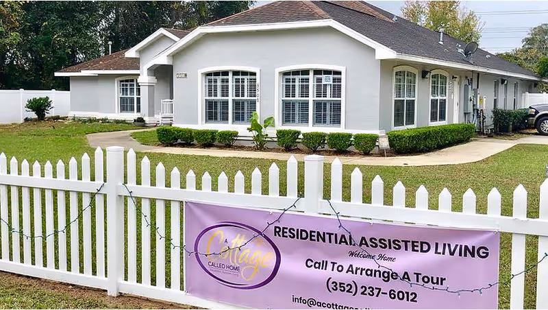 Exterior view of a single-story residential assisted living facility named 'A Cottage Called Home' with white walls, multiple windows, and a brown roof. A white picket fence surrounds the property, and a purple banner on the fence advertises residential assisted living and provides a phone number and email for arranging a tour.