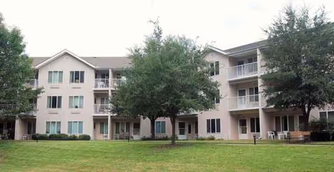 Beige senior living building with balconies, a manicured lawn, and trees.