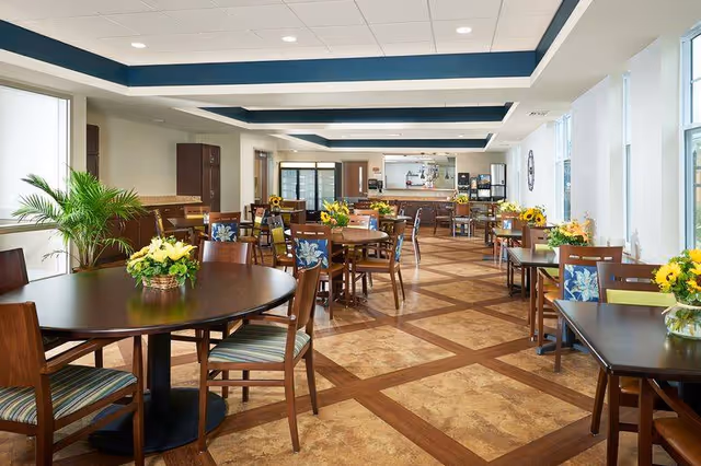 Bright dining room with round and square wooden tables and chairs, floral centerpieces, large windows, and a serving counter in the background.