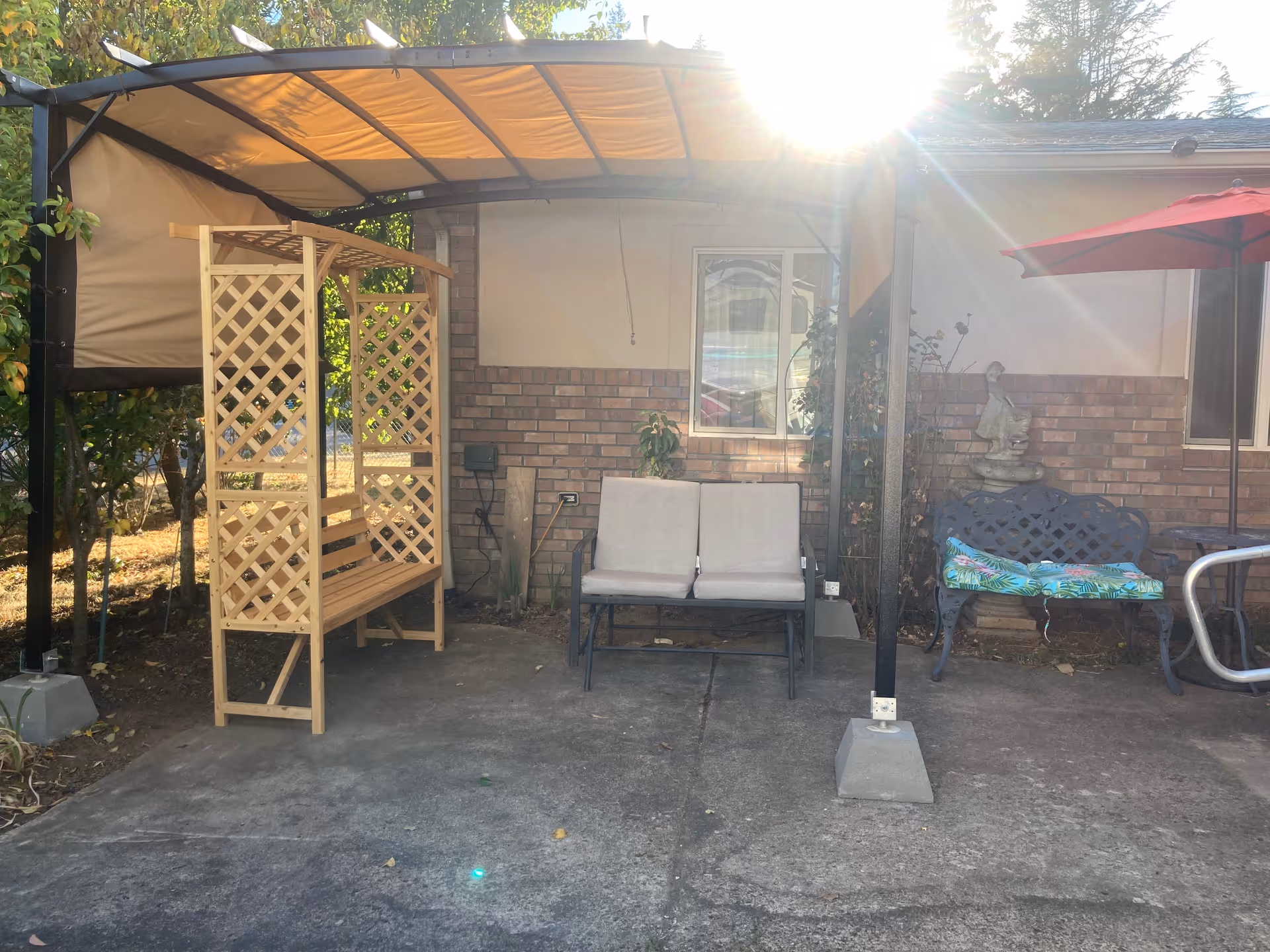 Outdoor patio area with a wooden lattice bench under a canopy, a two-seater cushioned bench, and a metal bench with a colorful cushion and a red umbrella. The background shows a brick wall with a window and some plants, with sunlight shining from the upper right corner.