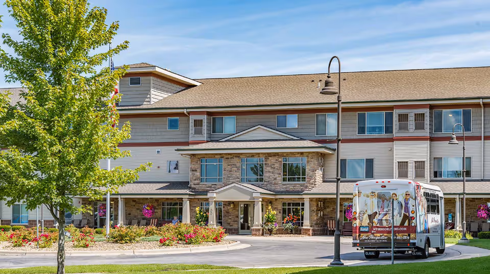 Exterior view of a senior living facility building with three stories, featuring a mix of stone and siding on the facade. There is a circular driveway in front with a small bus parked, decorated with images of seniors. The area is landscaped with green grass, a tree, flower beds, and hanging flower baskets. Two street lamps are visible near the driveway.