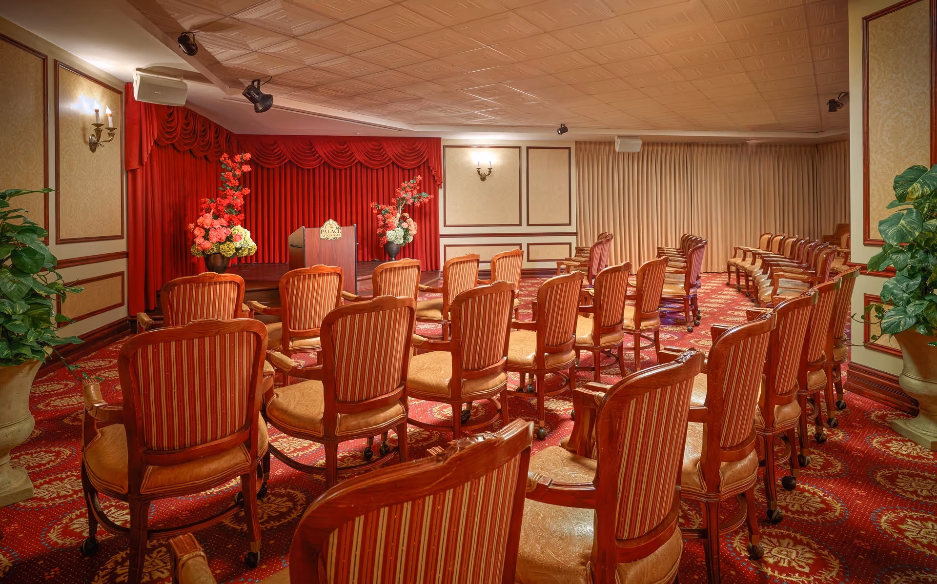 An elegant conference or presentation room with rows of wooden chairs upholstered in red and gold striped fabric facing a small stage with a podium. The stage backdrop is a rich red curtain with floral arrangements on either side. The room has ornate carpeting with red and gold patterns, beige walls with wood paneling, and potted plants on either side near the front.