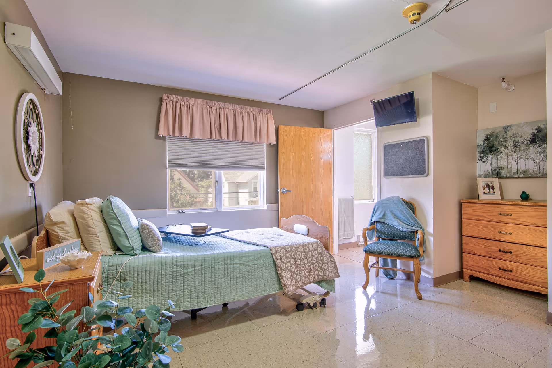 A sunlit, neatly furnished senior bedroom with a single bed, bedside table, armchair, dresser, and wall-mounted TV.