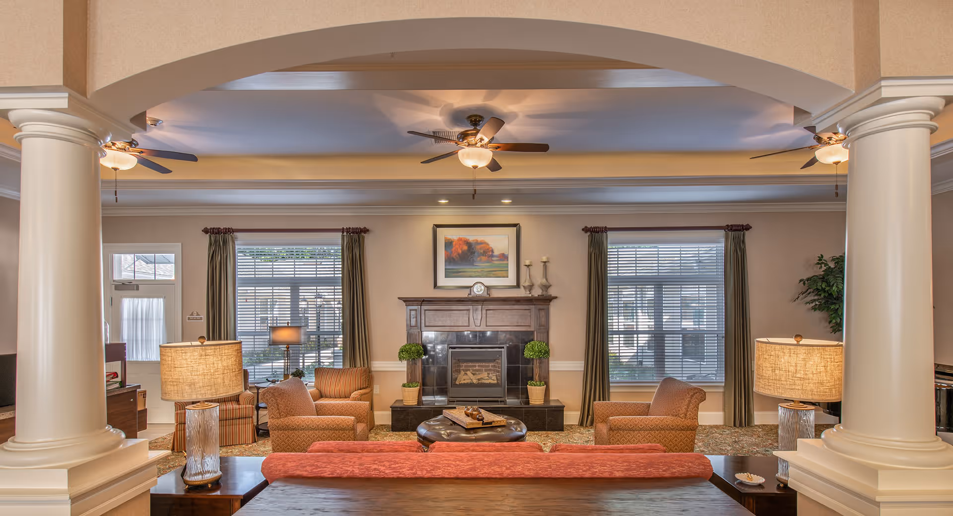 Elegant communal living room with columns, sofas and armchairs arranged around a central fireplace under ceiling fans.