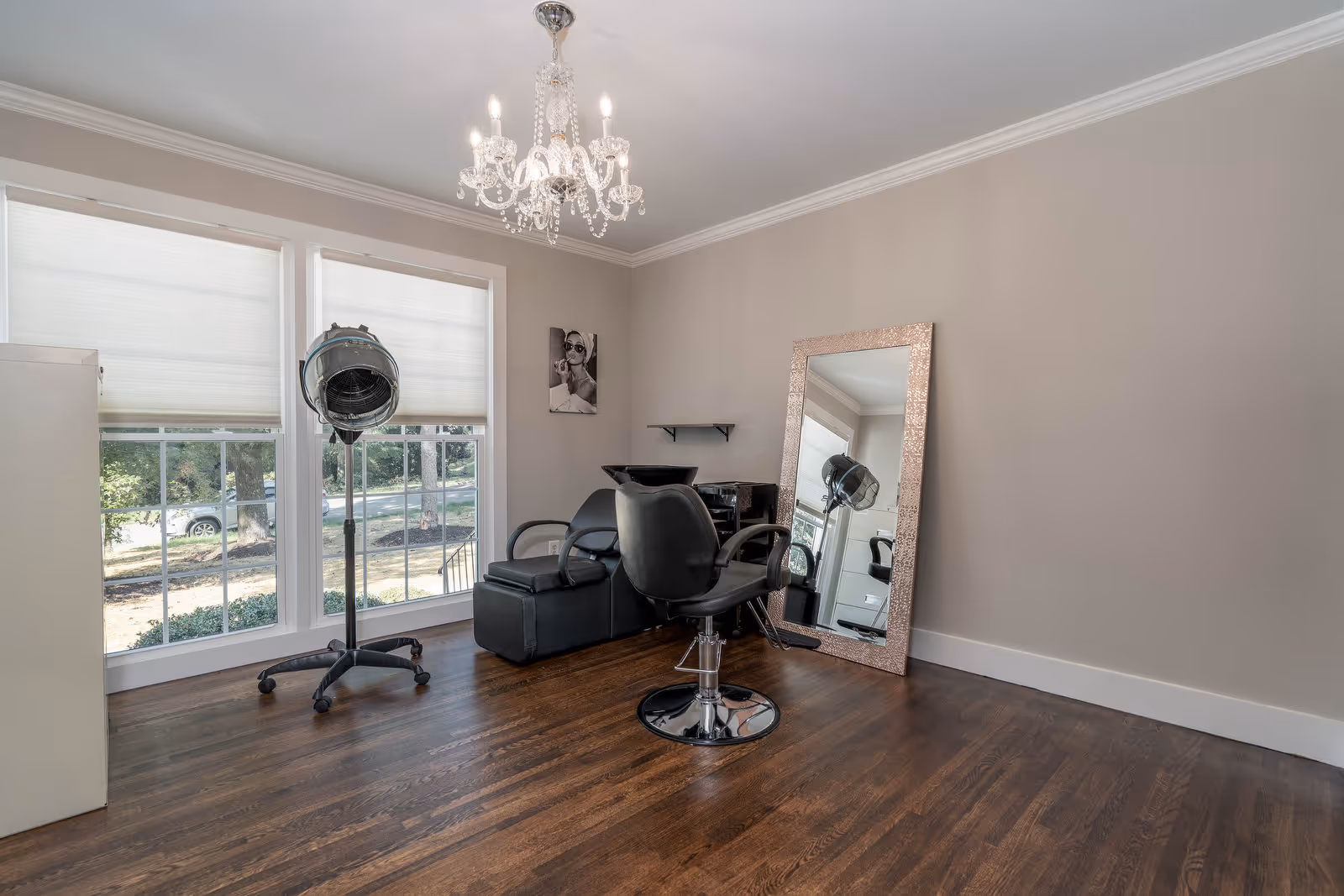 A small salon area with a black salon chair, a hair dryer on a stand, a large framed mirror leaning against the wall, and a black shampoo station. The room has large windows with white blinds, light gray walls, and dark hardwood floors. A chandelier hangs from the ceiling.