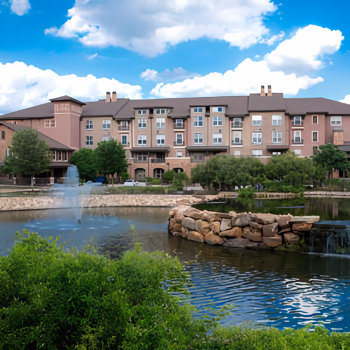 Exterior view of a large multi-story senior living facility building with multiple windows, surrounded by green trees and bushes. In the foreground, there is a pond with clear water, a stone wall, and a small waterfall. The sky is blue with scattered white clouds.