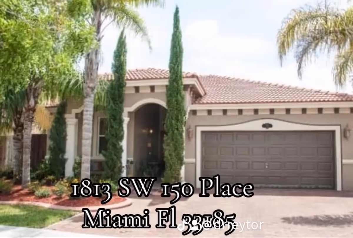 A single-story beige residence-style building with a red-tiled roof, arched entry, a two-car garage, and tall trees in the landscaped front yard.