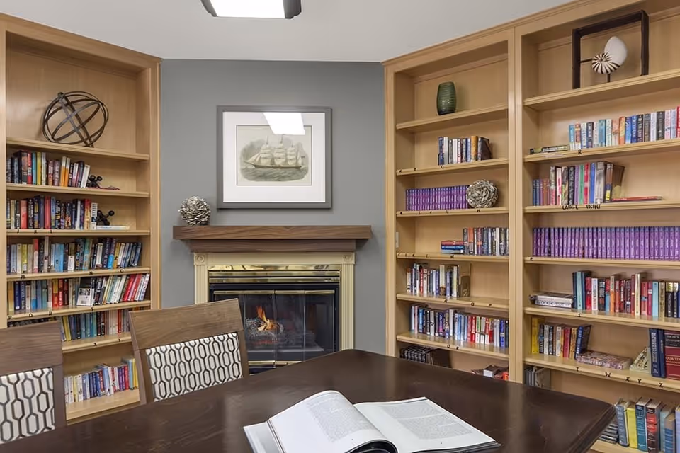 A cozy library room with built-in wooden bookshelves filled with books on either side of a fireplace with a lit fire. Above the fireplace hangs a framed picture of a ship. In front of the fireplace is a dark wooden table with an open book and two chairs with patterned cushions.