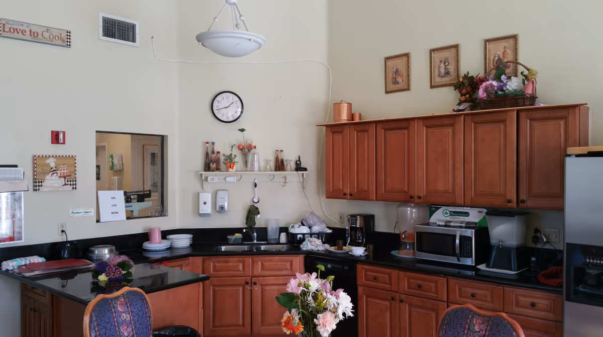 Interior view of a kitchen area with wooden cabinets, a black countertop, a microwave, a coffee maker, a water dispenser, and various decorative items including flowers and framed pictures on the wall. There is a clock on the wall and a sign that says 'Love to Cook'.
