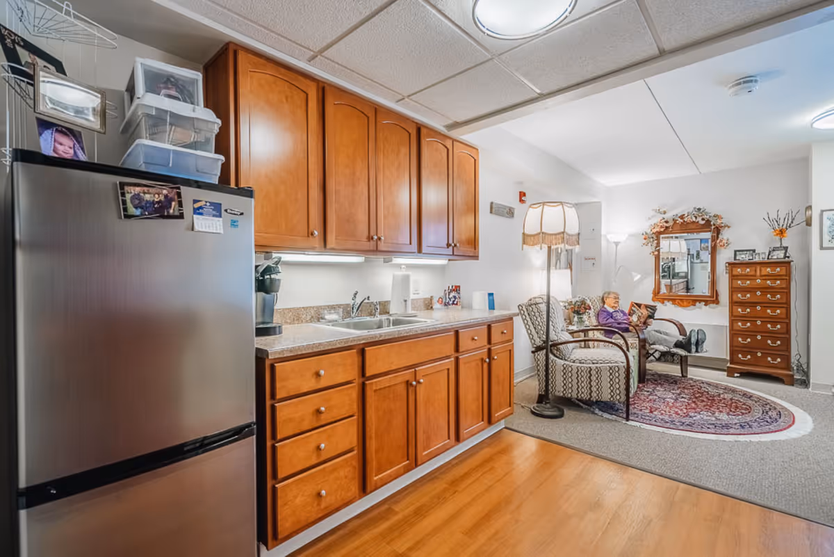 Interior view of a senior living facility room showing a kitchenette with wooden cabinets, a stainless steel refrigerator, and a countertop with a sink and coffee maker. In the adjacent sitting area, an elderly woman is seated in a patterned armchair reading a book, with a floor lamp, a decorative mirror, a chest of drawers, and a rug on the floor.