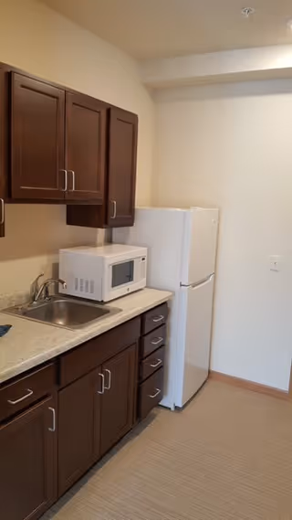 Small kitchen area with dark brown cabinets, a white microwave on the countertop, a stainless steel sink, and a white refrigerator against a plain wall.