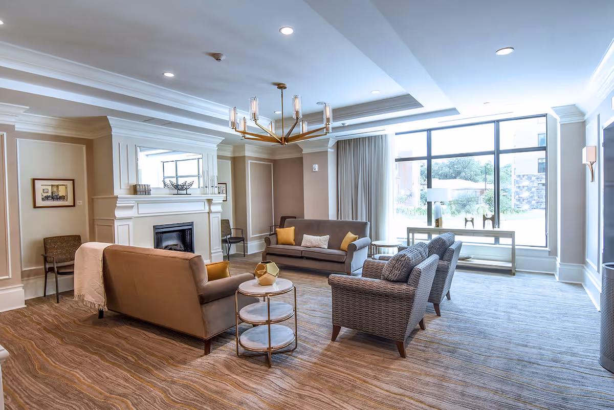 A bright and spacious living room with large windows letting in natural light. The room features two brown sofas, two patterned armchairs, a round side table with a decorative object, a fireplace with a large mirror above it, and a modern chandelier hanging from the ceiling. The walls are painted in neutral tones with white trim, and there is a console table with a lamp near the window.