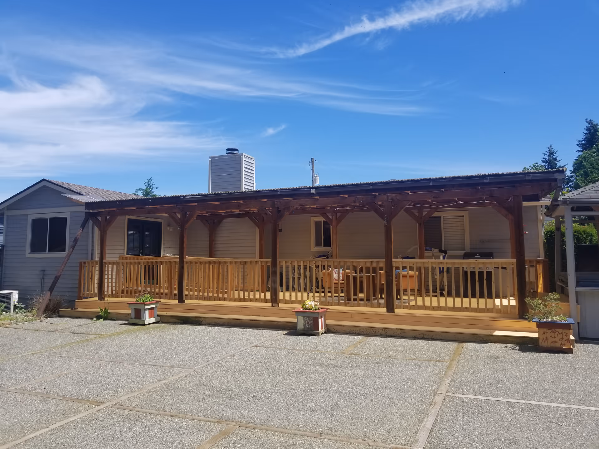 Exterior view of a single-story building with a wooden covered porch and railing. The porch has some outdoor furniture and potted plants. The sky is clear with some wispy clouds.