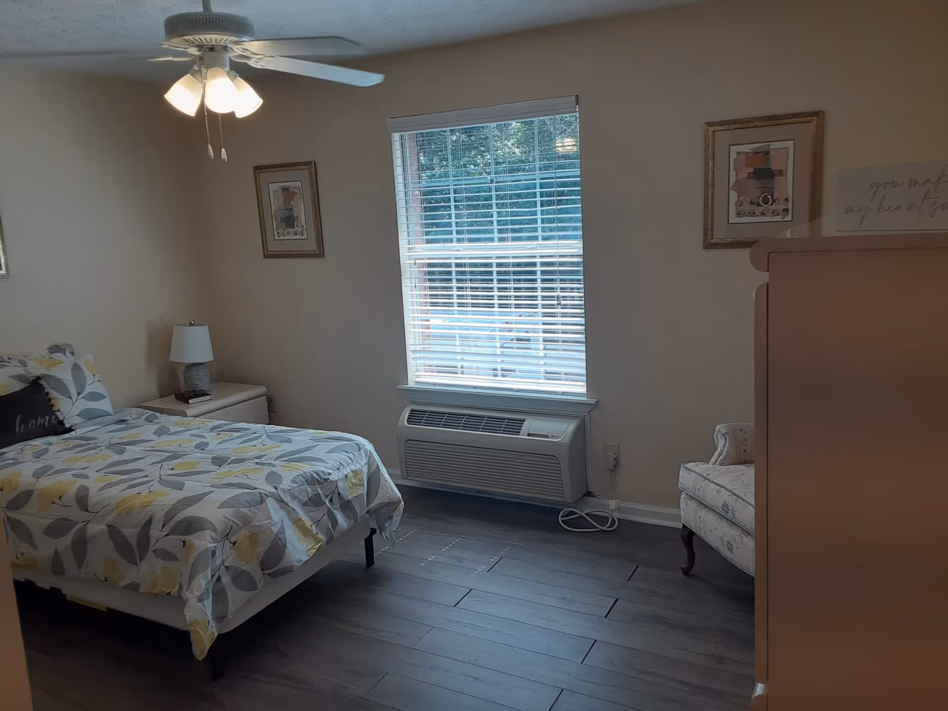 A bedroom with a bed covered in a floral patterned bedspread, a nightstand with a lamp, a ceiling fan with lights, a window with blinds, a cushioned armchair, and a dresser.