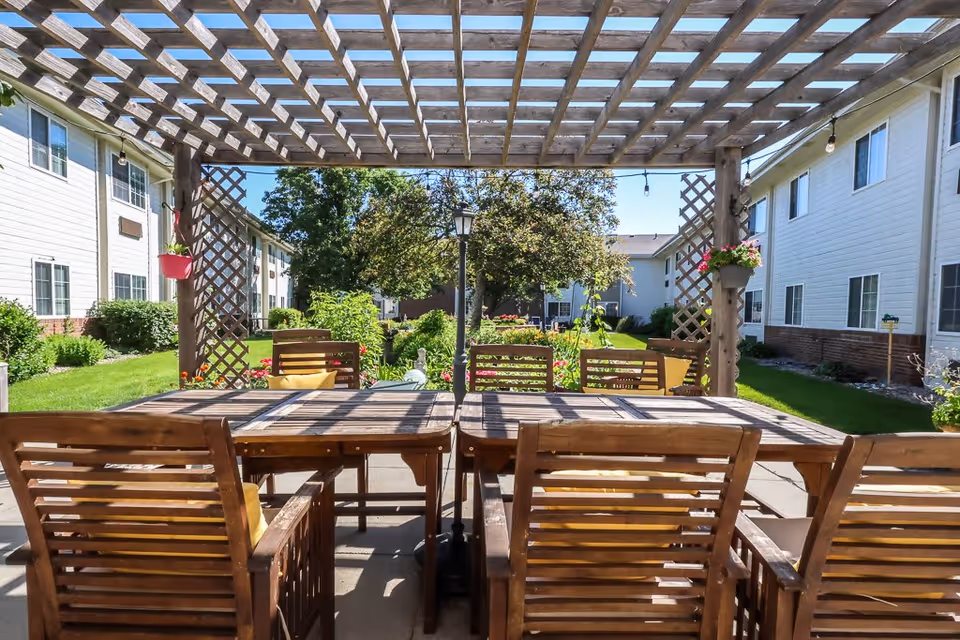 Outdoor seating area with wooden tables and chairs under a wooden pergola, surrounded by greenery and flowers, with two-story white buildings on either side and a clear blue sky.