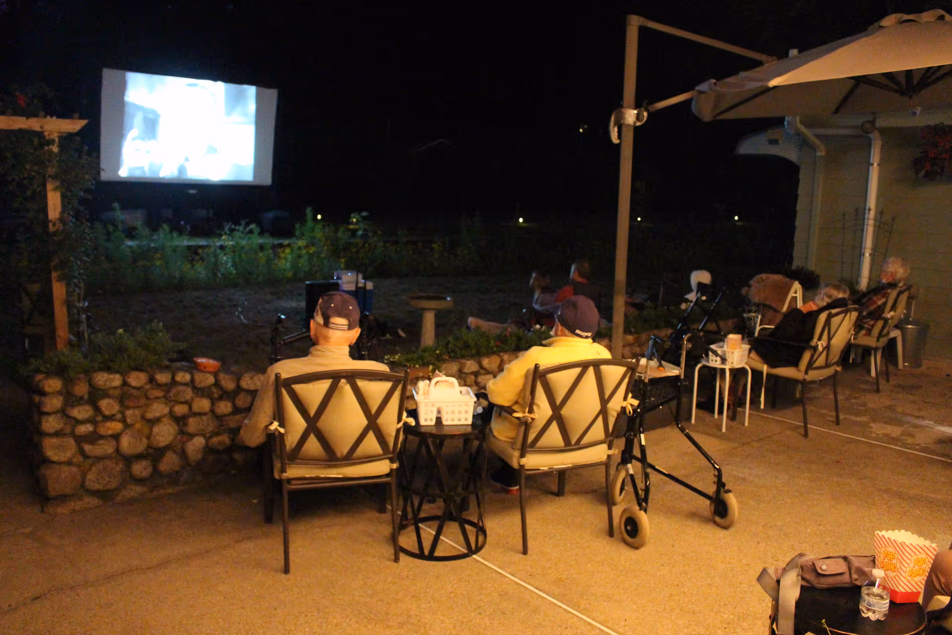 Seniors seated on patio chairs at night watching a movie projected on an outdoor screen, with walkers and refreshments nearby.