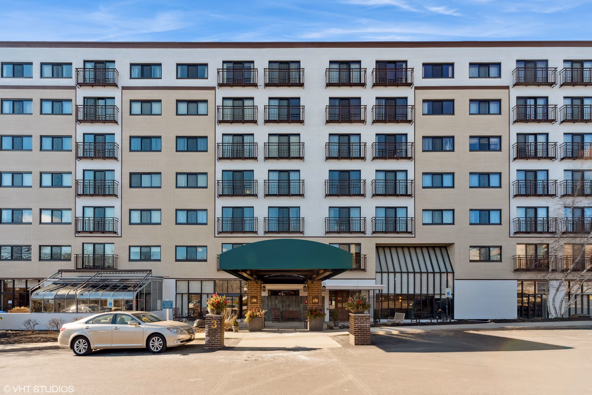 Front exterior view of a multi-story senior living facility with multiple windows and balconies. There is a green awning over the main entrance, flanked by two brick pillars with flower arrangements. A beige car is parked in front of the building.
